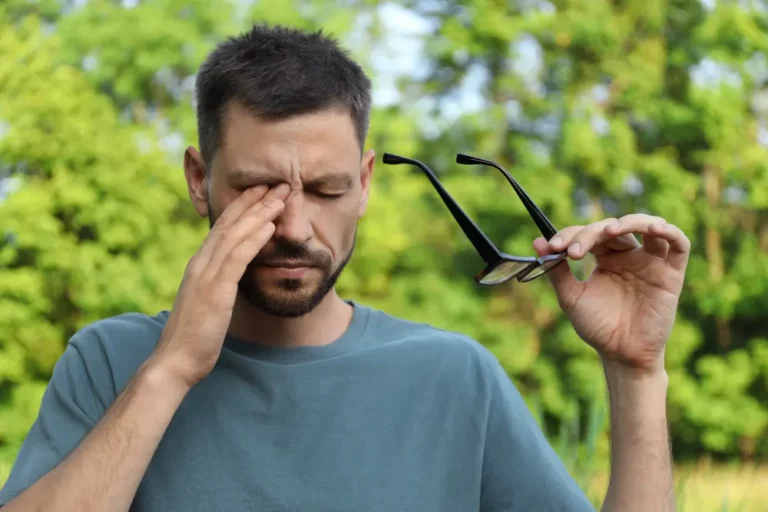 Mann draußen in der Natur fasst sich mit der Hand an sein rechtes Auge und hält seine Brille in der anderen Hand.